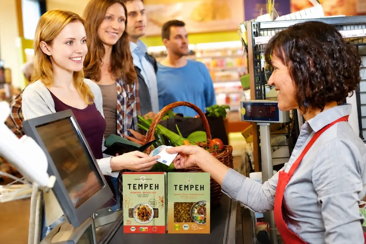 Kundin bezahlt an der Supermarktkasse, vor ihr stehen zwei Tempeh-Packungen auf dem Kassenband.