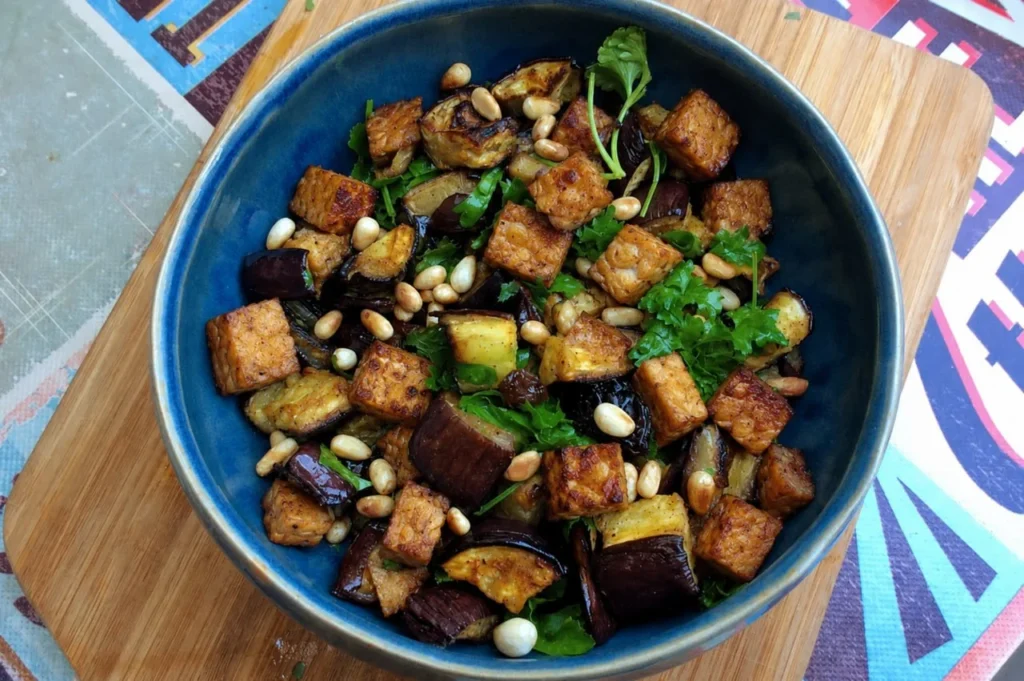 Knusprige Tempeh-Würfel mit gerösteter Aubergine, Pinienkernen und Petersilie in einer blauen Schale
